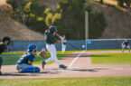 A Menlo Park Legends batter in a dark green jersey makes contact with a pitch during a preseason game, with the ball visible just off the bat. A blue-uniformed catcher and home plate umpire frame the play, and a fielder in the background readies for the ball near the 310-foot marker.