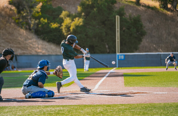 A Menlo Park Legends batter in a dark green jersey makes contact with a pitch during a preseason game, with the ball visible just off the bat. A blue-uniformed catcher and home plate umpire frame the play, and a fielder in the background readies for the ball near the 310-foot marker.